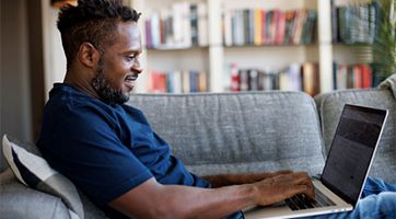 Man sitting sideways on a couch, typing on a laptop in his lap